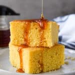 Two stacked pieces of cornbread on a white plate being drizzled with honey. A towel and a small bottle of honey is in the background.