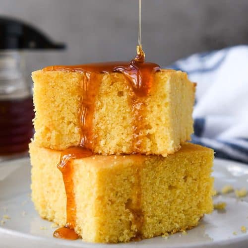 Two stacked pieces of cornbread on a white plate being drizzled with honey. A towel and a small bottle of honey is in the background.