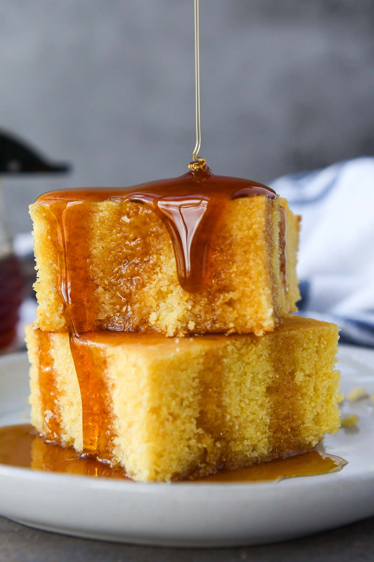 Two stacked pieces of cornbread on a white plate being drizzled with honey. A towel and a small bottle of honey is in the background.