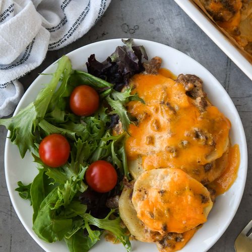 Above view of a plateful of ground beef casserole and a side salad. The casserole dish, a fork, and a white striped towel surround the plate.