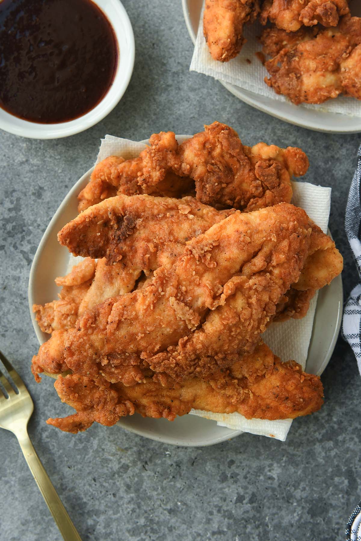 Above view of a plate piled high with chicken tenders with a bowl of bbq sauce, a second plate of tenders, a fork, and black plaid towel.