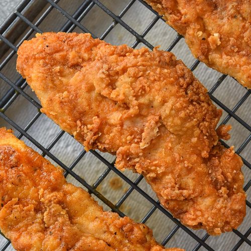 Above view of three chicken tenders draining on a rack over a paper towel lined baking sheet.