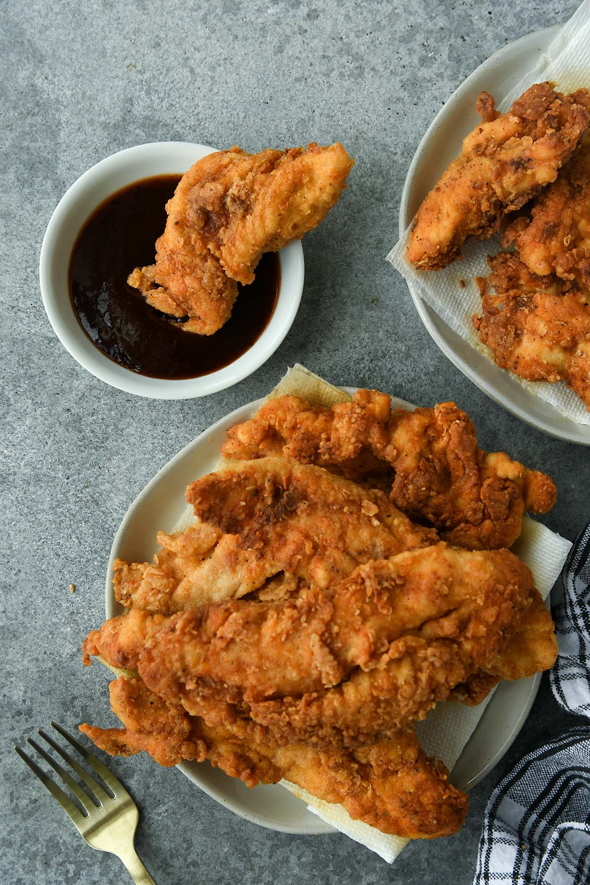 Above view of a plate piled high with chicken tenders with a tender in a bowl of bbq sauce, a second plate of tenders, a fork, and black plaid towel.