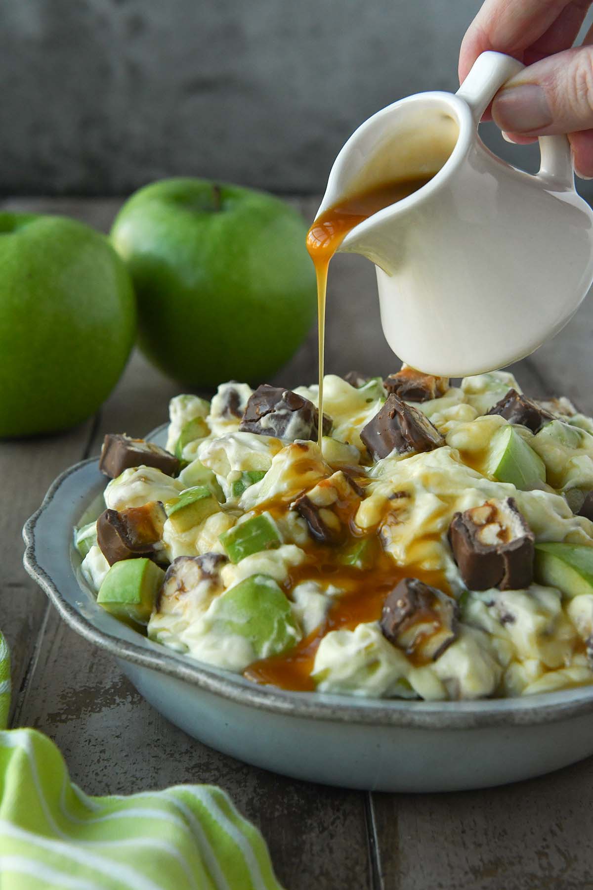A small white pitcher pouring caramel over a bowl of apple caramel snickers salad with two apples in the background.
