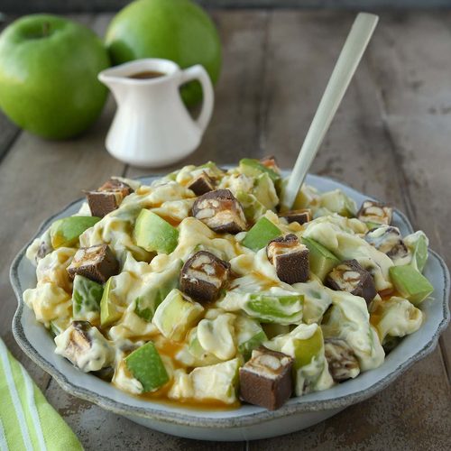 A bowl of snickers salad with two green apples in the background and a small pitcher. A green towel in the foreground.