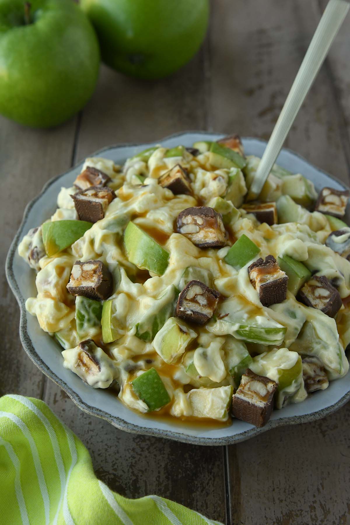 A bowl of snickers salad with two green apples in the background and a small pitcher. A green towel in the foreground.