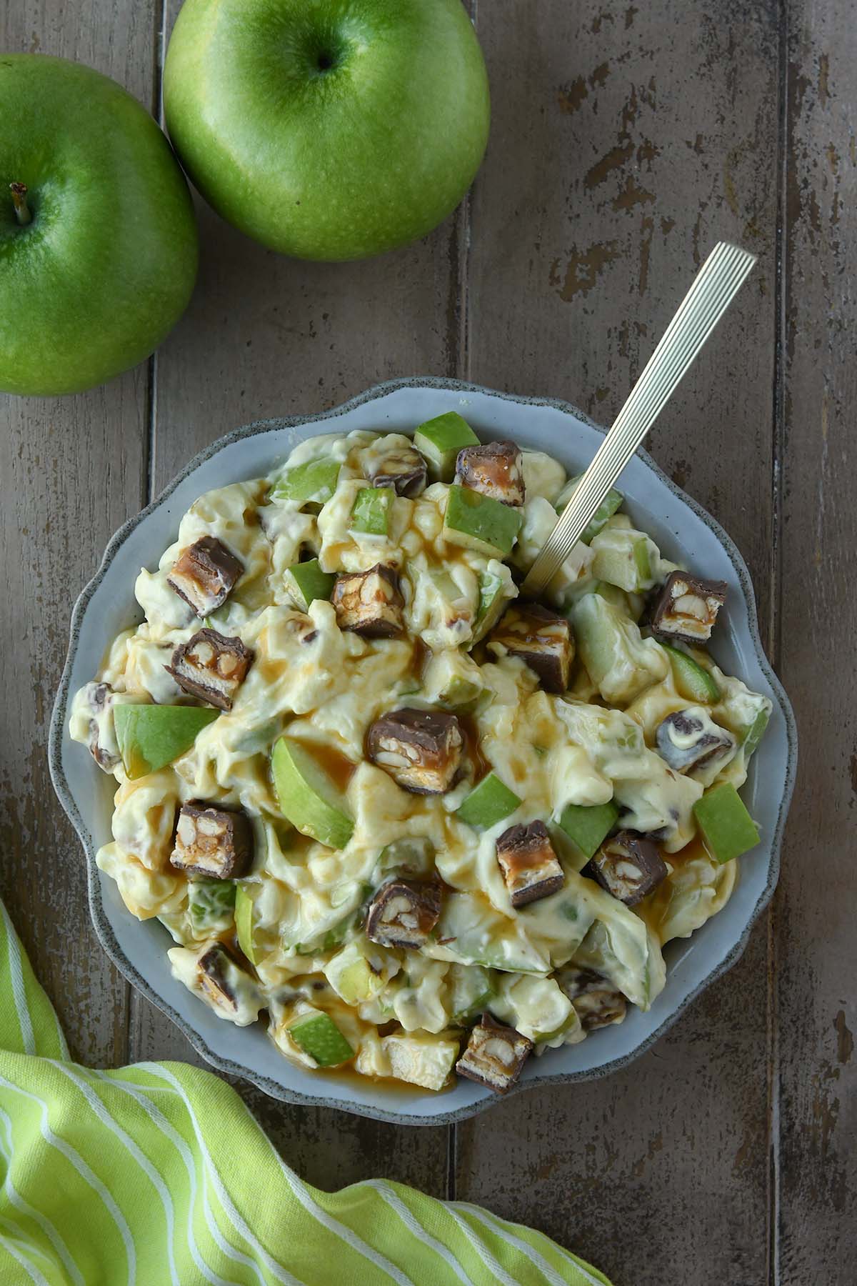 Above view of a bowl of snickers salad with two green apples and a green towel.