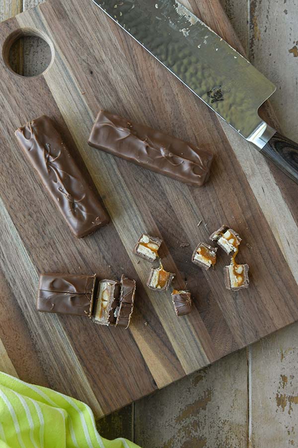 Step one is cutting the Snickers up into bite-sized pieces on a cutting board with a knife and a green striped towel.