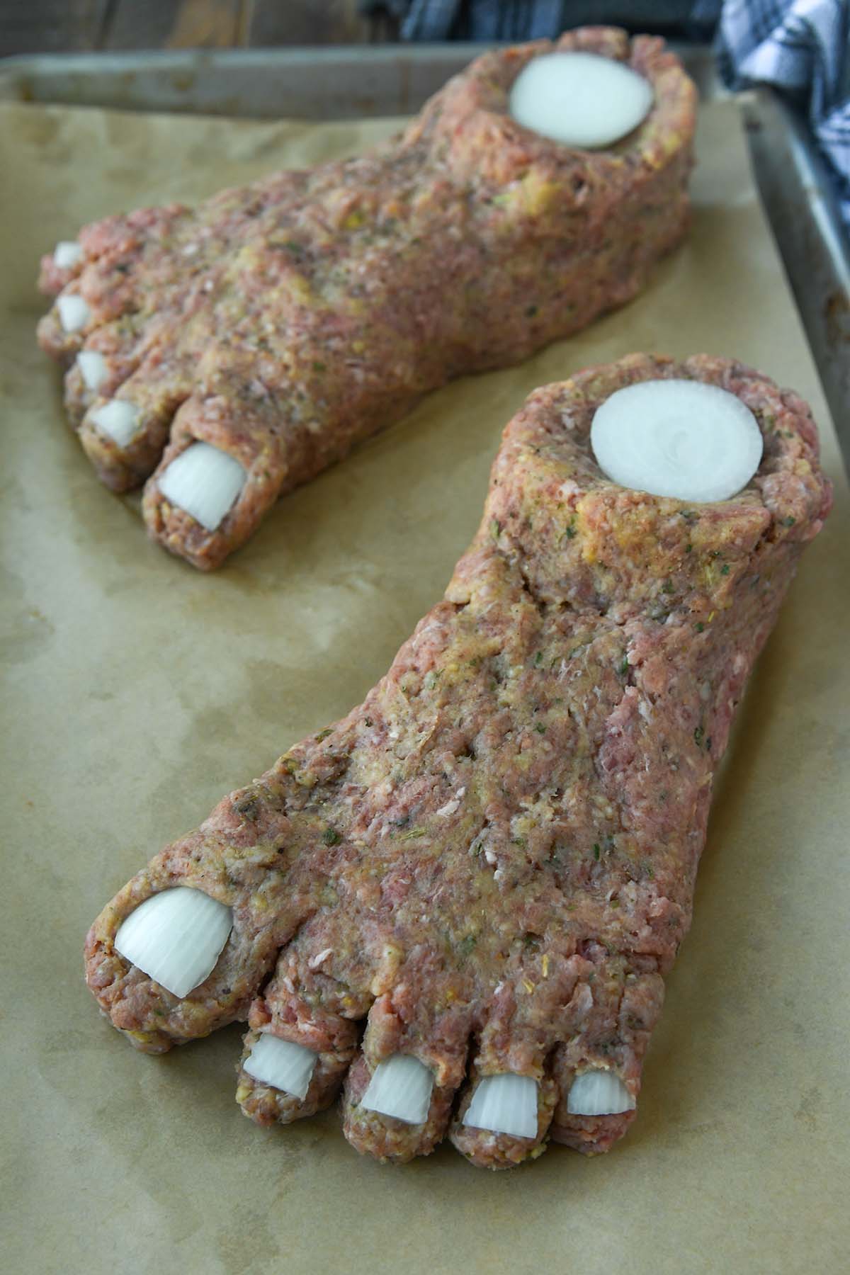 Up close for feet formed out of meatloaf mixture and the onion toenails added on a baking sheet with a towel in the background.