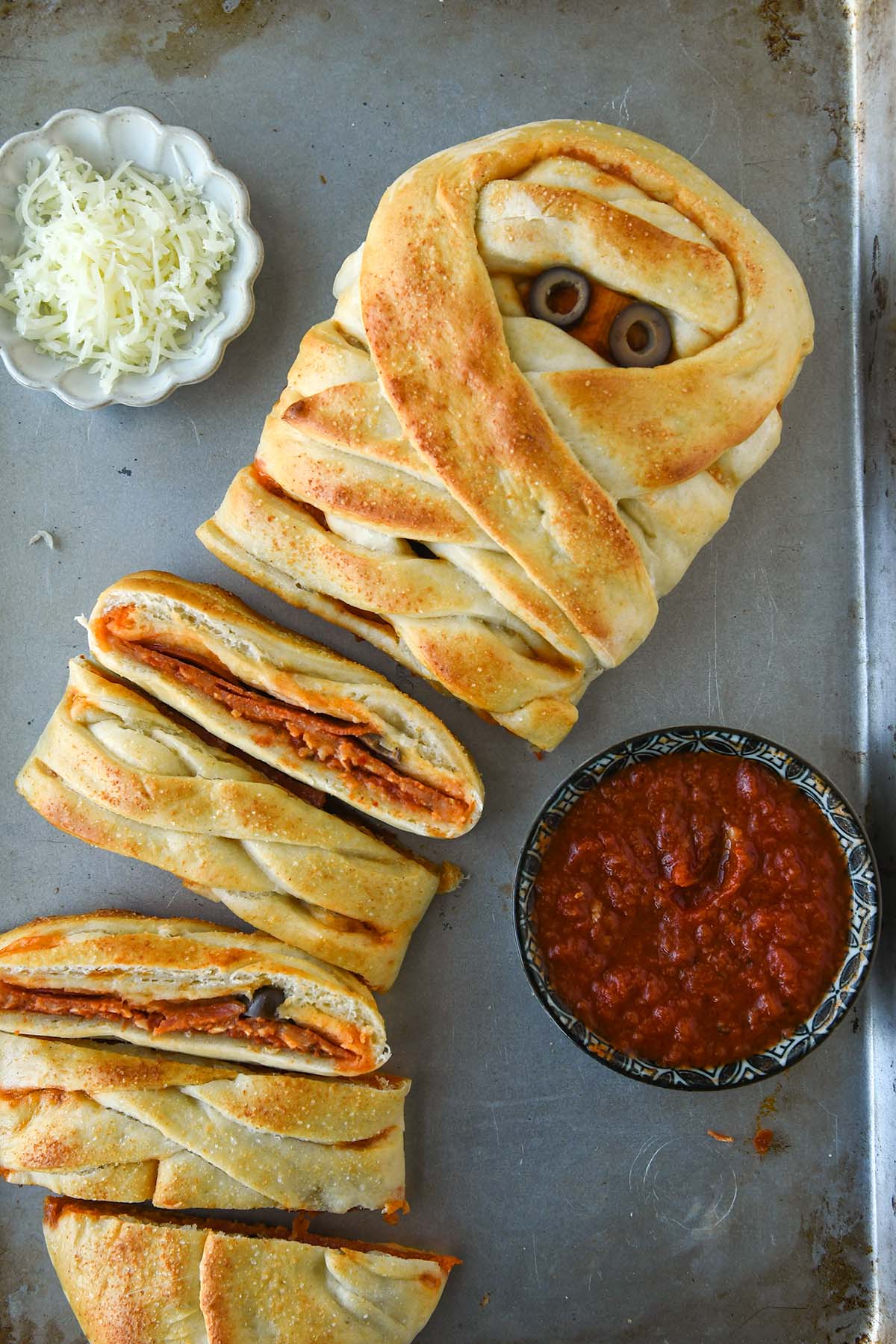 Above view of the pizza mummy sliced up and served with a side of sauce and a small bowl of cheese.