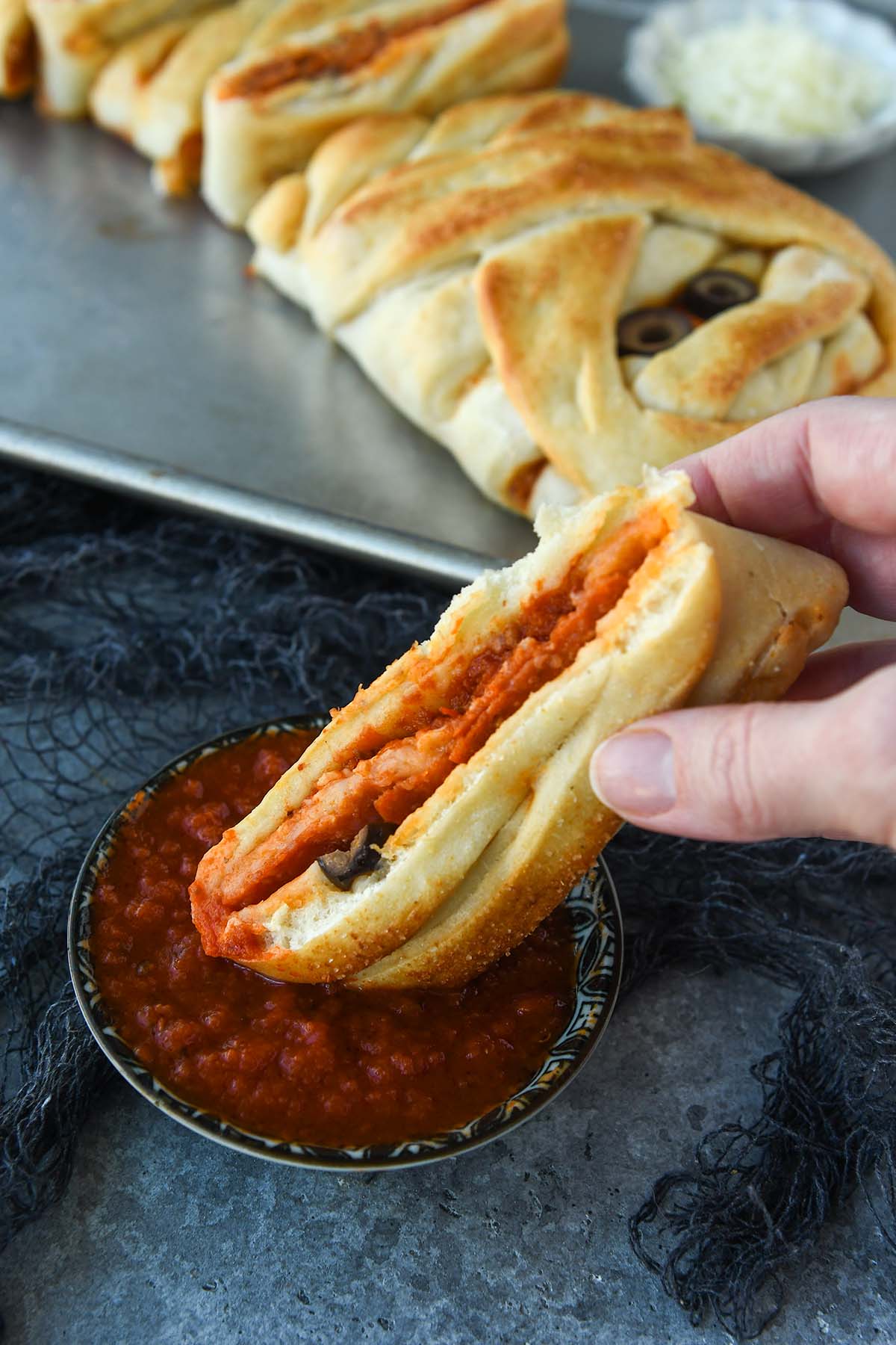A slice of pizza mummy being dipped into marinara sauce with the mummy braid in the background.