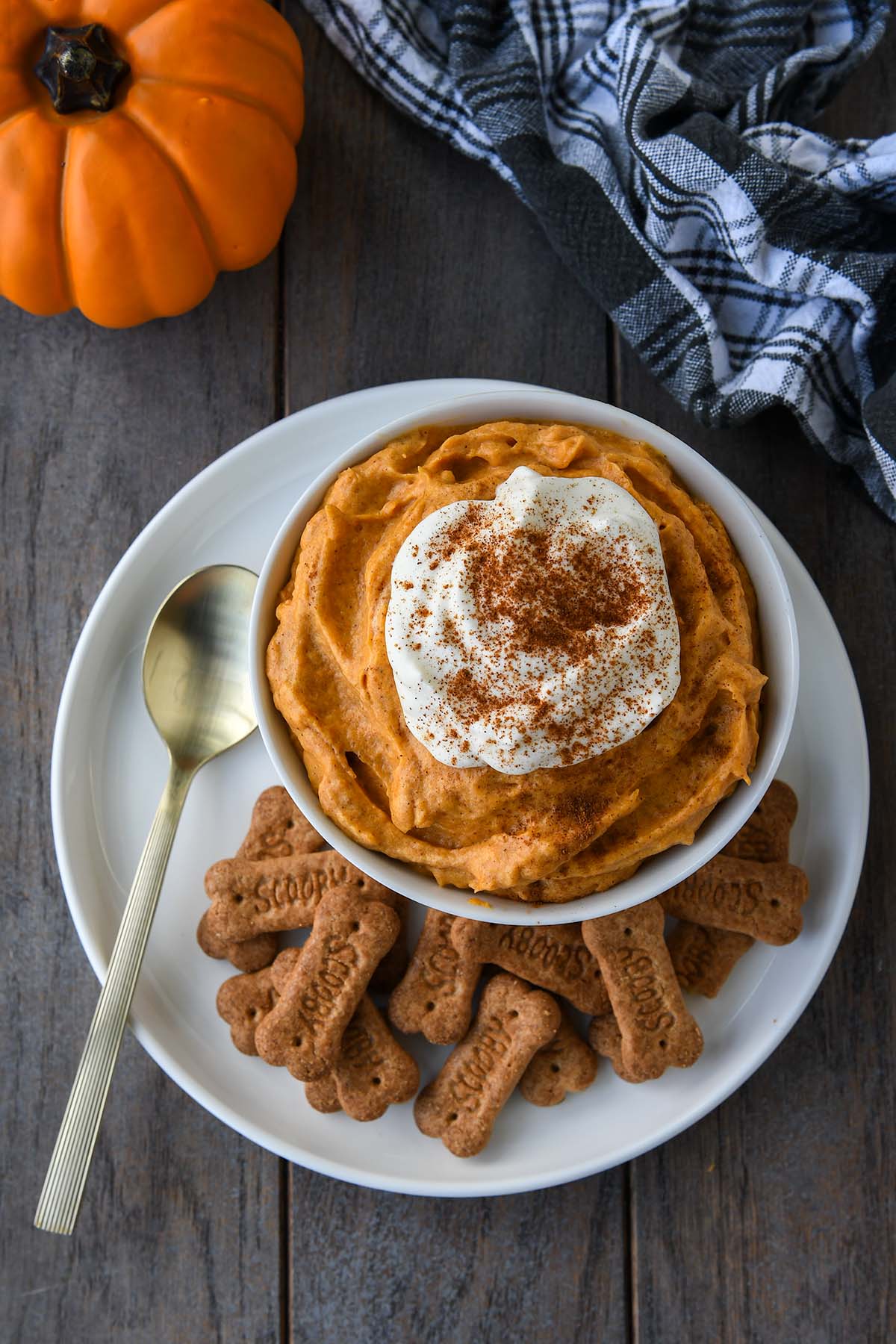 Above view of a bowl of pumpkin fluff surrounded by graham cracker sticks with a spoon. A small pumpkins and a black plaid towel in the background.