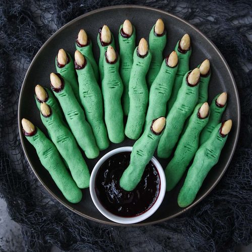 Above view of a dark plate of witch finger cookies with a small bowl of raspberry jam.