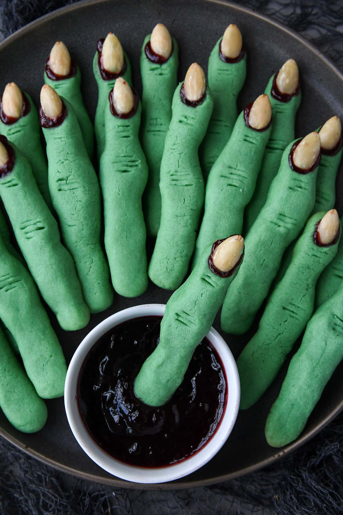 Above view of a dark plate of witch finger cookies with a small bowl of raspberry jam.